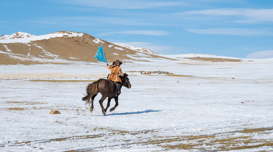 Kazakh rider holding flag galloping across frozen Mongolian steppe.